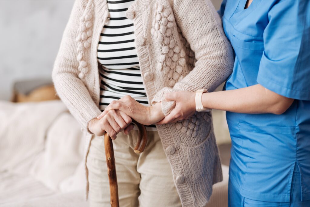 Caregiver in scrubs steadying an older woman who is walking with a cane